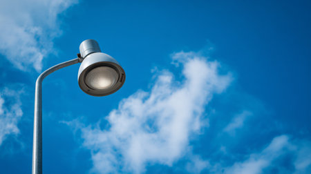 A sleek street lamp stands tall against a bright blue sky filled with soft white clouds. This daytime image captures urban lighting and architectural design in harmony.の素材