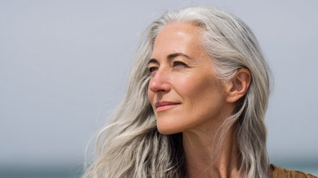 Elegant mature woman with long silver hair enjoys a moment of tranquility as she gazes off into the distance, surrounded by a beautiful natural seascape.の素材
