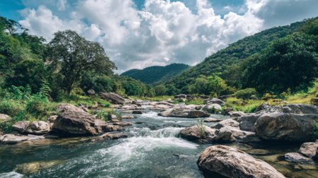 A peaceful river flows through a vibrant green landscape, surrounded by rolling hills and dramatic clouds, showcasing the tranquility and beauty of unspoiled nature.の素材