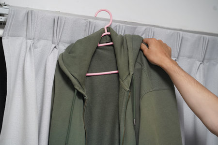 A hand holds a green hoodie on a pink hanger, showcasing a cozy style in a minimalist indoor setting. The soft fabric backdrop adds a serene touch to the scene.の写真素材