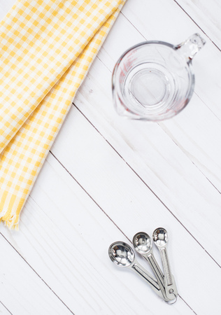 A glass measuring cup and metal teaspoons sit on a wood top next to yellow dish towelの写真素材