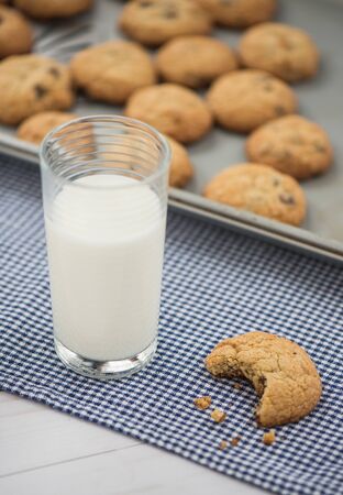 A glass of milk and a chocolate chip cookie with a bite taken out of it sit on a blue gingham napkin with a cookie sheet in the backgroundの写真素材