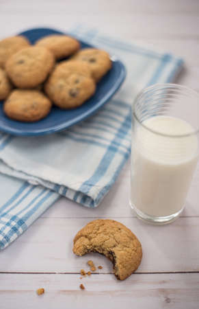 A glass of milk and a cookie with a bite taken out of it next to a blue plaid napkin with a plate of cookies in the backgroundの写真素材