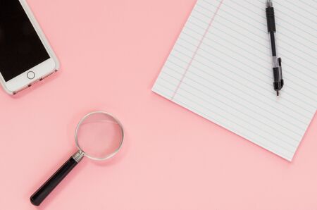 A smart phone, writing pad, pen and magnifying glass on a pink surfaceの写真素材