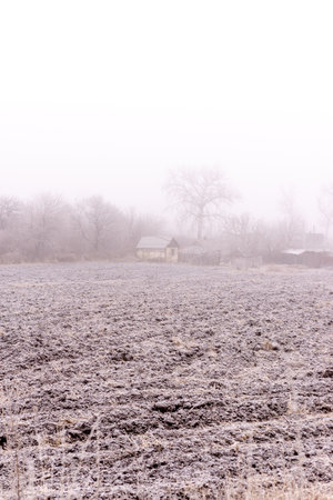 A serene winter scene featuring a frost-covered plowed field and a small rustic house in the background. Surrounded by bare trees and faint mist, the image captures the calm and beauty of rural life during the colder months.の写真素材
