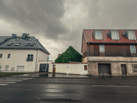 Quiet, overcast day on a rainy village street. The wet asphalt and dramatic sky contrast with the old, worn residential buildings and a green, ivy-covered hedge.の写真素材