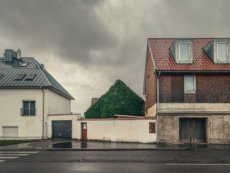 Quiet, overcast day on a rainy village street. The wet asphalt and dramatic sky contrast with the old, worn residential buildings and a green, ivy-covered hedge.の写真素材