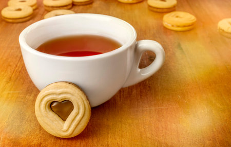 A white cup of tea and a butter cookie with a heart-shaped chocolate filling, on a dark wooden texture, more blurred cookies and bokehs in the background, with copyspaceの写真素材