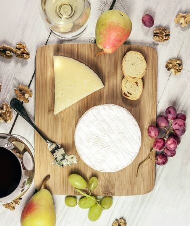 A photo of a tasting with glasses of wine, red and white, different types of cheese, bread, nuts, pear, grapes, on a wooden boardの写真素材