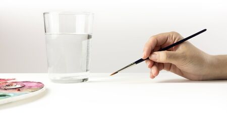 A photo of a hand holding a watercolor brush, with a palette, a water glass in the blurred background, and a blank sheet of paper with plenty of copyspaceの写真素材