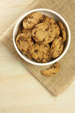 A photo of chocolate chips cookies in a bowl, shot from above on a burlap and light wooden background texture, with copyspaceの写真素材