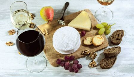 A photo of a tasting with glasses of wine, red and white, different types of cheese, bread, nuts, pear, grapes, on a wooden boardの写真素材