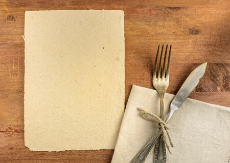 An overhead photo of a piece of parchment for copyspace, with a vintage fork and knife on a textile napkin. A restaurant menu or special offer banner design templateの写真素材