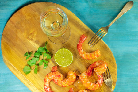 An overhead photo of a glass of white wine with cooked shrimps, a slice of lime, cilantro leaves, salt flakes, and a place for text, on a vibrant blue backgroundの写真素材