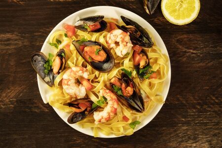 A photo of a ready to eat seafood pasta dish with mussels and shrimps, shot from above on a dark wooden rustic background texture, with a lemon half and some shells, with a place for textの写真素材