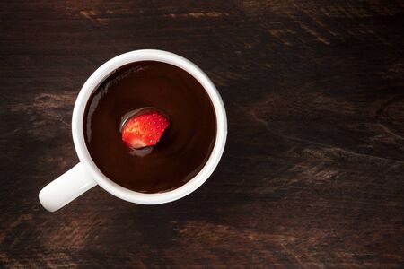 An overhead photo of a strawberry in a cup of dark chocolate, shot from above on a rustic wooden texture with a place for textの写真素材