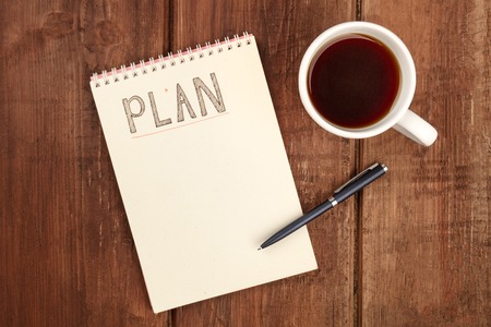 A photo of a spiral notebook with the word Plan, with a cup of coffee and a pen, shot from the top on a dark rustic wooden desk with a place for textの写真素材