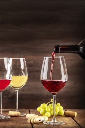 Red wine poured into a glass from a bottle, on a dark background, with other wineglasses, a place for text and grapes in the blurred background. Design template for a tasting invitationの写真素材