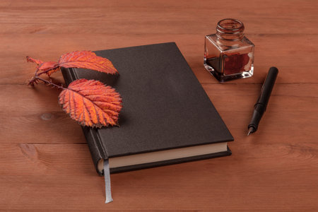 Autumn Poetry. A photo of a black book with a thorny branch with vibrant leaves, an ink well, and a nib pen, on a dark wooden table with copy spaceの写真素材
