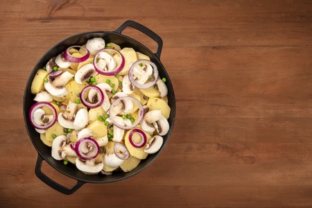 A photo of a braiser with raw vegetables, potatoes, onions, green peas, and mushrooms, shot from the top on a dark rustic wooden background wih a place for textの写真素材