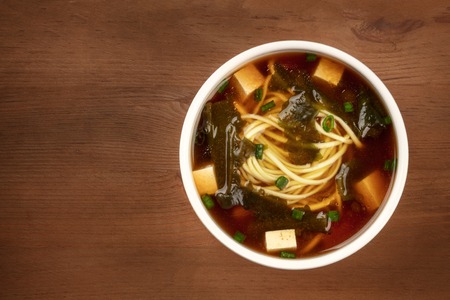 A bowl of miso shiru soup with tofu, scallions, noodles, and wakame seaweed, shot from the top on a rustic wooden background with copy spaceの写真素材