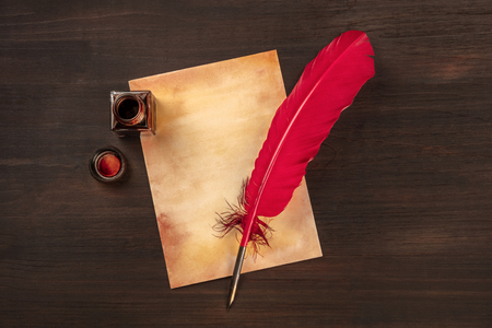 A vintage background with a vibrant red quill on a piece of old paper, with an ink well, on a dark wooden background with copy spaceの写真素材
