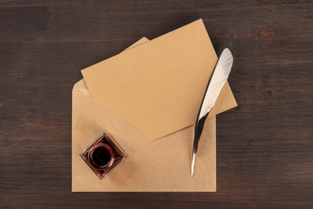 An ink well with a brown craft card in an envelope, with a quill pen and copy space, on a dark wooden background. A mockup for an invitation or greeting cardの写真素材