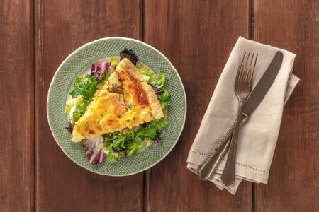 A slice of French quiche with salmon, with green salad leaves, shot from above on a dark rustic wooden backgroundの写真素材
