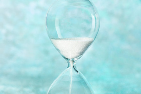 Time is running out concept. A close-up of an hourglass with sand falling through, on a teal blue...