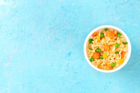 Instant noodles with vegetables in a plastic cup, overhead shot on a blue background with a place for textの写真素材