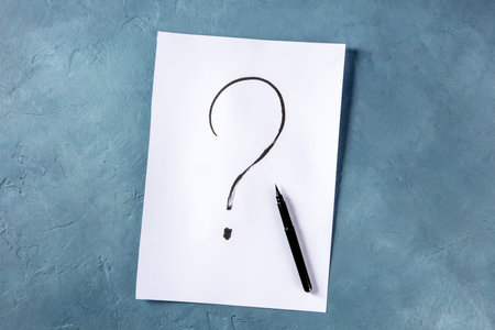 Question mark, written in ink on a piece of standard office paper, shot from the top on a blue...