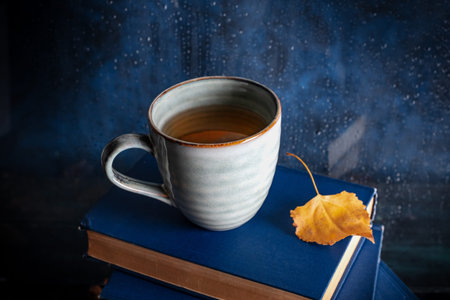 Autumn reading. A cup of tea and an autumn leaf on a stack of books in front of a window with rain behind itの写真素材