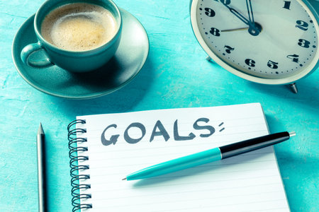 Goal setting. A notepad with the handwritten word Goals, with a pen, an alarm clock, and a cup of...