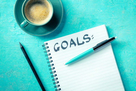 Goal setting. A notebook with the handwritten word Goals, with a cup of coffee, a pen, and a pencil, overhead flat lay shot on a vibrant blue background. The concept of writing a list or a planの写真素材
