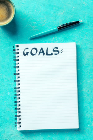 Goals, handwritten word in a notepad with a ballpoint pen and a cup of coffee, shot from the top on a vibrant blue background. The concept of goal setting, writing a list or a planの写真素材