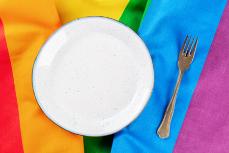Gay dinner concept. A place at a restaurant table with a plate and a fork, overhead flat lay shot on the LGBT rainbow flag textureの写真素材