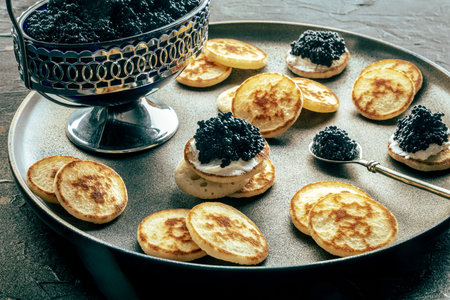 Blinis with black caviar and cream cheese, on a festive dish on a black background, mini pancakes, an elegant appetizerの写真素材