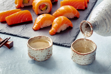 Sake poured into ochoko, traditional cup, with salmon nigiri, sashimi, and roll in the backgroundの写真素材