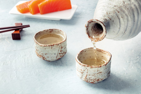 Sake poured into ochoko, traditional cup, with salmon sashimi in the background and chopsticks. Traditional Japanese food and drink, with copy spaceの写真素材