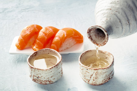 Sake poured into ochoko, traditional cup, with salmon nigiri in the background, traditional Japanese food and drinkの写真素材