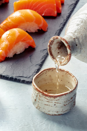 Sake poured into ochoko, traditional cup, with salmon nigiri in the background. Traditional Japanese drinkの写真素材