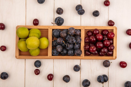 top view of fresh fruits such as green cherry plumred cherries and dark purple sloes on a wooden divided tray on a white wooden background