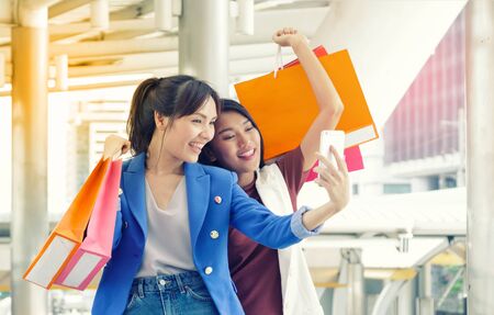 Two Asian woman taking selfie with smartphone while holding shopping bags in the shopping mallの写真素材