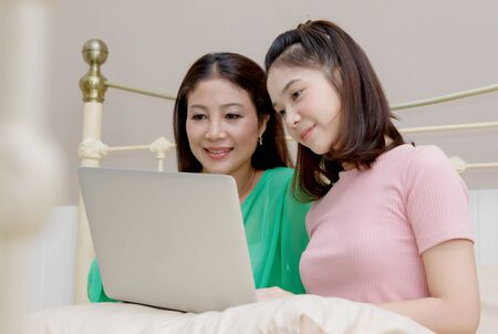 Asian mother and her teenage daughter are using computer while sitting on bedの写真素材