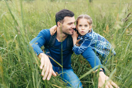 Father with daughter having walk on green grass, enjoying every moment of being together. Little playful kid embracing her father who is paving way in high grass, looking at her daughter with loveの写真素材