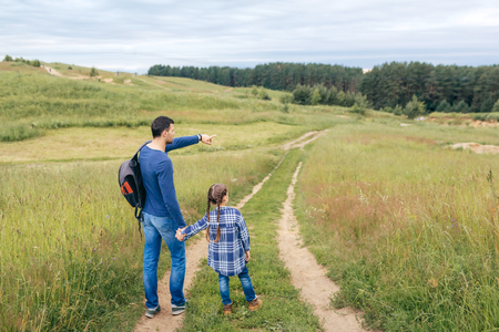 Horizontal portrait of little girl with long pigtails wearing checkered shirt keeping her hand in daddy s hand while walking with him on path, looking into distance on green forest. Family, natureの写真素材