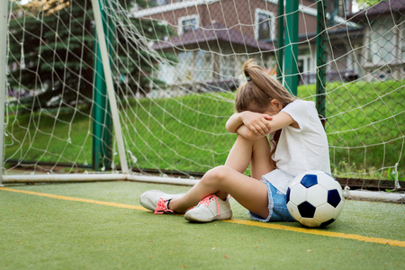 Crying ltittle girl who is playing football, hiding her face while being upset to miss goal in gates. Pretty kid wearing white t-shirt, jean shorts and sport shoes, playing football. Children, sportの写真素材