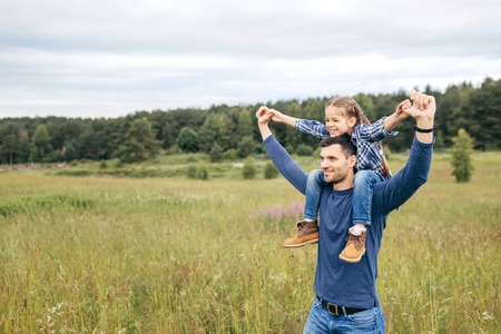 Happy cute daughter piggybacking on her father to see beautiful nature while having stroll together. Handsome young dad and his little child playing on nature. Father showing something to his daughterの写真素材