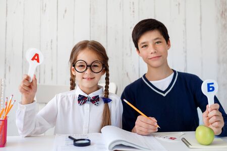 Schoolboy and schoolgirl sitting together, learning alphabet, raising stencils with letters, writing in their copy books, reading books with magnifying glass. Schoolchildren preparing for lessonsの写真素材