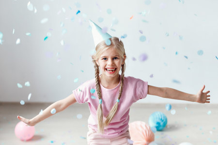 Joyful smiling blonde female kid with two pigtails, wears festive hat, tries to catch flying papers...